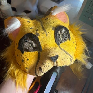 Op de afbeelding: Een geel, op een katachtig dier ge&iuml;nspireerd masker met zwarte stippen, lange gele imitatiebont plukken aan de zijkanten en witte oorplukjes. Het masker heeft zwart omrande gaas-oogopeningen, een kleine zwarte driehoekige neus en een textuurafwerking.