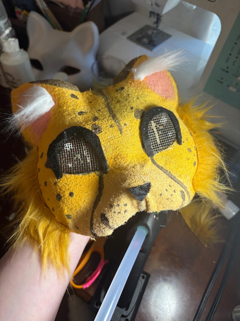 Op de afbeelding: Een geel, op een katachtig dier ge&iuml;nspireerd masker met zwarte stippen, lange gele imitatiebont plukken aan de zijkanten en witte oorplukjes. Het masker heeft zwart omrande gaas-oogopeningen, een kleine zwarte driehoekige neus en een textuurafwerking.