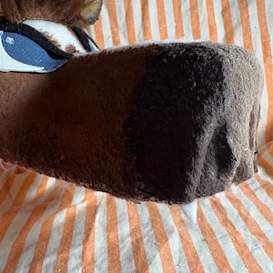Op de afbeelding: Een bruin pluche dierenkopmasker met een donkerbruine snuit, een pluk bruin imitatiebont aan de bovenkant en blauw-witte oogdetails. Het masker heeft een textuurrijk oppervlak en is voorzien van zichtbare naden langs de zijkanten en rond de ogen.