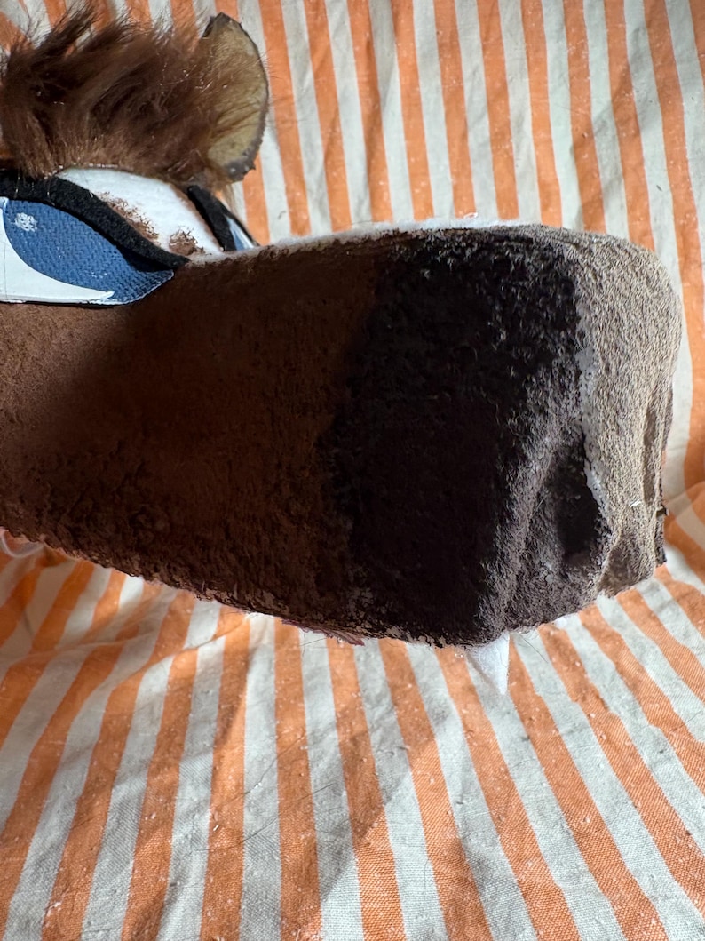 Op de afbeelding: Een bruin pluche dierenkopmasker met een donkerbruine snuit, een pluk bruin imitatiebont aan de bovenkant en blauw-witte oogdetails. Het masker heeft een textuurrijk oppervlak en is voorzien van zichtbare naden langs de zijkanten en rond de ogen.