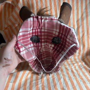 Op de afbeelding: Een handgemaakt dierenmasker met een rood-witte ruitjesvoering, twee donkerbruine rechtopstaande oren, een kleine pluk bruin haar aan de bovenkant en zwarte mesh oogbedekkingen. Een dunne witte elastische band is bevestigd om het masker vast te zetten.