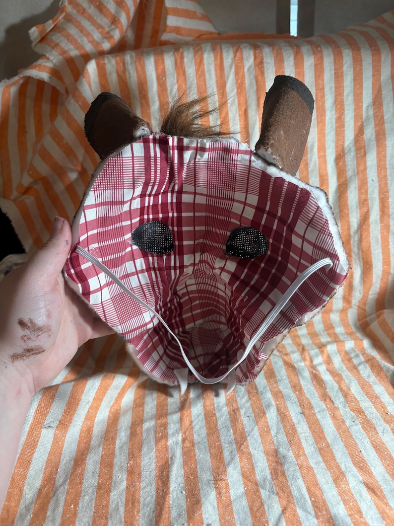 Op de afbeelding: Een handgemaakt dierenmasker met een rood-witte ruitjesvoering, twee donkerbruine rechtopstaande oren, een kleine pluk bruin haar aan de bovenkant en zwarte mesh oogbedekkingen. Een dunne witte elastische band is bevestigd om het masker vast te zetten.