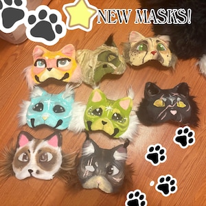 Op de afbeelding: Acht katachtige maskers met diverse geschilderde ontwerpen, kleuren en accenten van imitatiebont. De maskers tonen verschillende oogkleuren, patronen en uitdrukkingen. De tekst NEW MASKS! is bovenaan zichtbaar, vergezeld door pootafdruk-iconen.