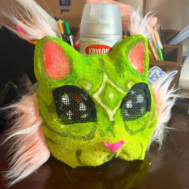 Op de afbeelding: Een limoengroen kattenmasker met roze imitatiebontplukjes op de oren en wangen, zwarte gaasbedekking voor de ogen, een kleine roze neus en een lichtgroen ruitpatroon op het voorhoofd. Het masker heeft een textuur en een ronde katachtige vorm.