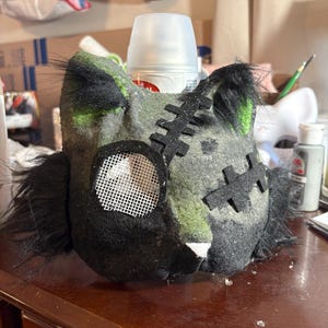 Op de afbeelding: Een handgemaakt kattenmasker van donkergrijs en zwart vilt met groene accenten. Het masker heeft zwart imitatiebont aan de zijkanten, een wit gaasje voor &eacute;&eacute;n oog en zwarte vilten stikdetails op het voorhoofd en de wang. Het heeft spitse oren.