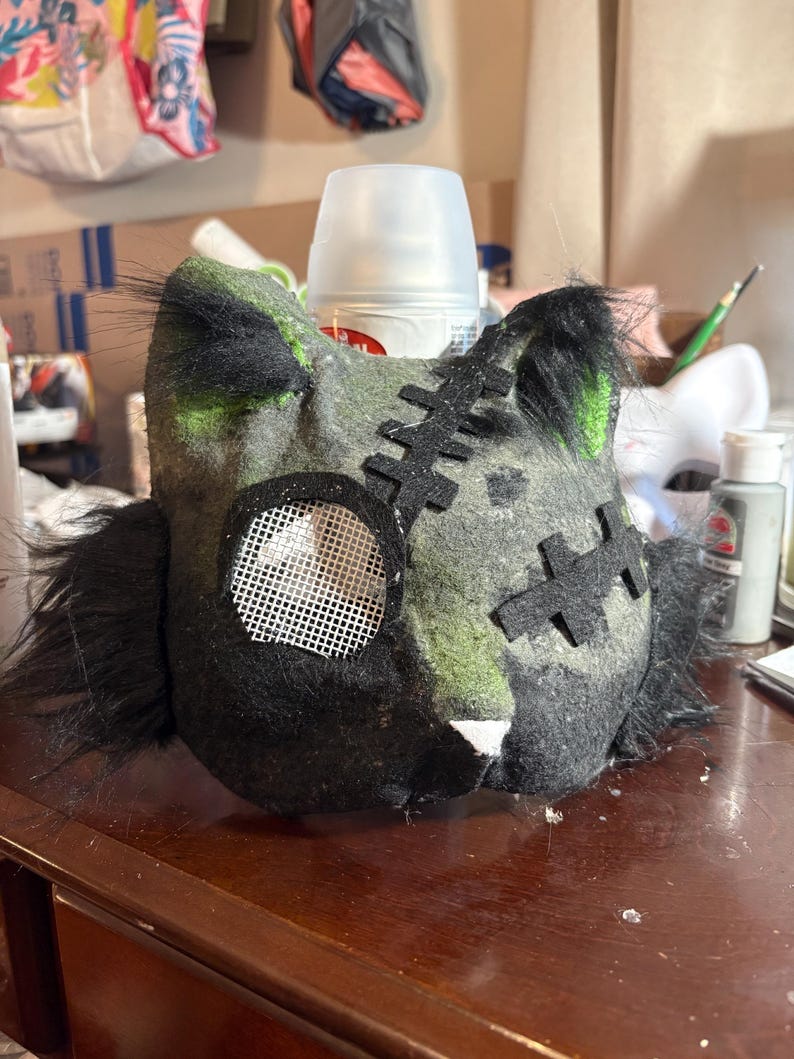Op de afbeelding: Een handgemaakt kattenmasker van donkergrijs en zwart vilt met groene accenten. Het masker heeft zwart imitatiebont aan de zijkanten, een wit gaasje voor &eacute;&eacute;n oog en zwarte vilten stikdetails op het voorhoofd en de wang. Het heeft spitse oren.