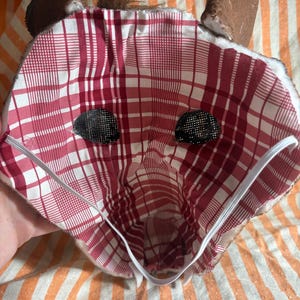 Op de afbeelding: Een stoffen dierenmasker met een rood-witte voering met ruitpatroon, twee donkere mesh-oogopeningen en een witte elastische band. De buitenkant van het masker is bruin met bruine oren en een klein plukje bruin bont aan de bovenkant in het midden.