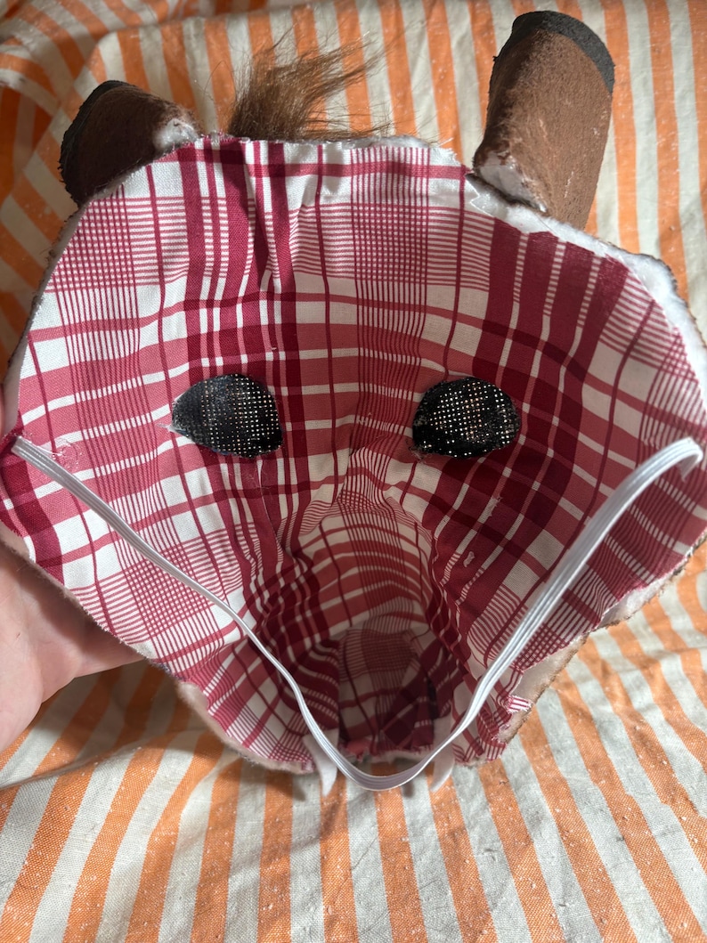 Op de afbeelding: Een stoffen dierenmasker met een rood-witte voering met ruitpatroon, twee donkere mesh-oogopeningen en een witte elastische band. De buitenkant van het masker is bruin met bruine oren en een klein plukje bruin bont aan de bovenkant in het midden.