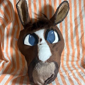 Op de afbeelding: Een bruin paardenmasker met een witte bles in het midden van het gezicht, donkerbruine vachtaccenten en grote blauwe ogen met witte omlijningen. Het masker heeft rechtopstaande oren en een pluk bruin haar op het voorhoofd met een textuur.