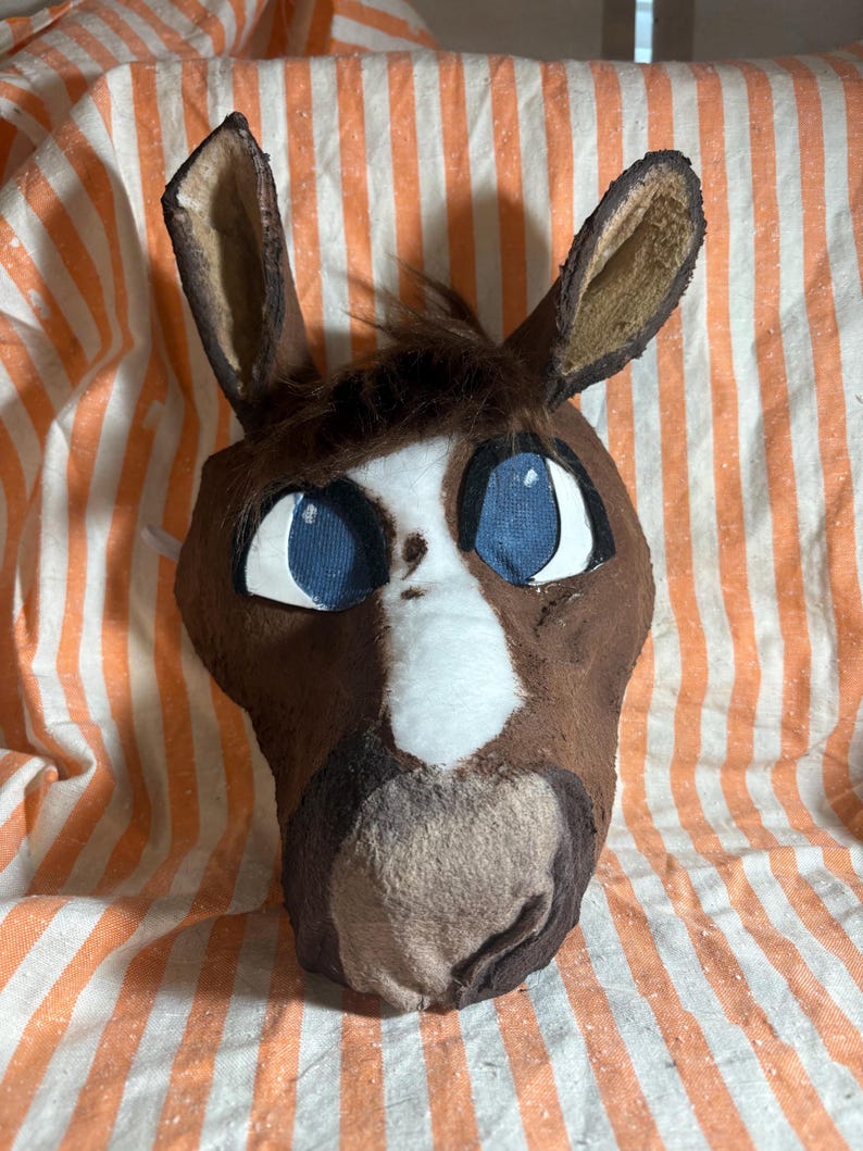 Op de afbeelding: Een bruin paardenmasker met een witte bles in het midden van het gezicht, donkerbruine vachtaccenten en grote blauwe ogen met witte omlijningen. Het masker heeft rechtopstaande oren en een pluk bruin haar op het voorhoofd met een textuur.