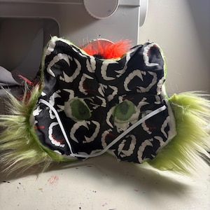 Op de afbeelding: Een pluizig groen masker met een zwarte stoffen voering met een wit herhalend tandpatroon. Het masker heeft twee groene mesh oogopeningen, een feloranje pluk imitatiebont aan de bovenkant en een dunne witte elastische band voor bevestiging.