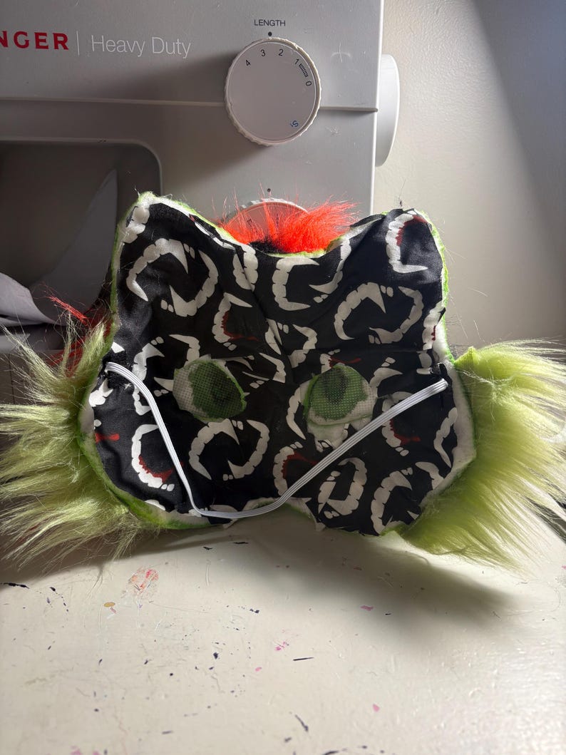 Op de afbeelding: Een pluizig groen masker met een zwarte stoffen voering met een wit herhalend tandpatroon. Het masker heeft twee groene mesh oogopeningen, een feloranje pluk imitatiebont aan de bovenkant en een dunne witte elastische band voor bevestiging.