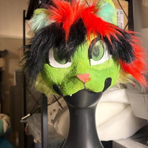 Op de afbeelding: Een op een kat ge&iuml;nspireerd masker met een felgroen, getextureerd gezicht, een kleine roze neus en zwarte markeringen. Het hoofddeksel is versierd met lange, felrode en zwarte plukken imitatiebont en heeft grote, witomrande mesh-oogopeningen.