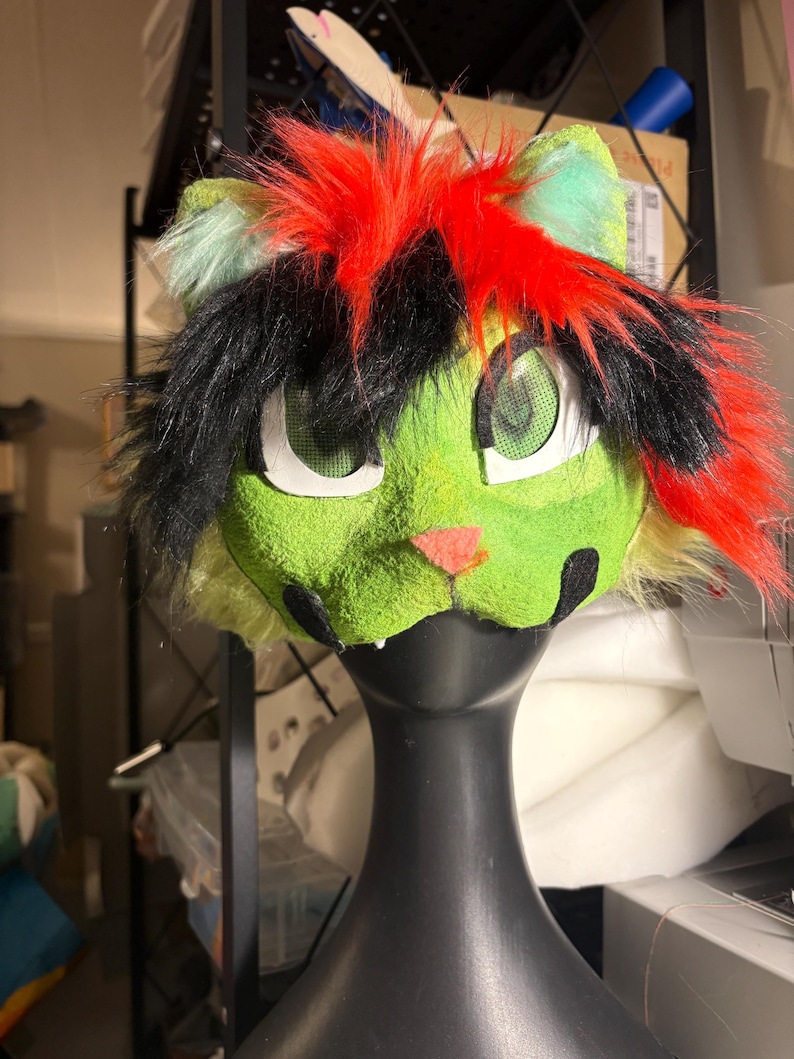 Op de afbeelding: Een op een kat ge&iuml;nspireerd masker met een felgroen, getextureerd gezicht, een kleine roze neus en zwarte markeringen. Het hoofddeksel is versierd met lange, felrode en zwarte plukken imitatiebont en heeft grote, witomrande mesh-oogopeningen.