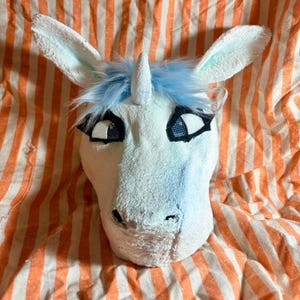Op de afbeelding: Een wit eenhoornmasker met een centrale hoorn, lichtblauw imitatiebont en grote ogen met zwarte omlijningen en gaasdetails. Het masker heeft twee puntige oren en een getextureerd oppervlak, ontworpen om het hoofd te bedekken.