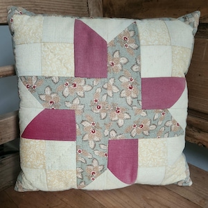May include: A square throw pillow featuring a patchwork design with a central star motif. The pattern combines solid dusty rose fabric, a light cream fabric with a subtle tonal print, and a sage green fabric decorated with a floral pattern.