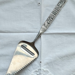 May include: A silver cheese slicer with a decorative handle featuring engraved figures and text. The blade has a serrated edge and a slotted design for slicing. The handle's design includes a figure with a bow and arrow.