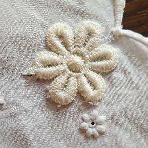 Op de afbeelding: Een wit textiel met een grote, verhoogde, cr&egrave;mekleurige geborduurde bloem met zes bloemblaadjes en een kleinere, platte, witte geborduurde bloem met een centraal oogje. De rand van de stof heeft een geschulpte, gestikte boord.