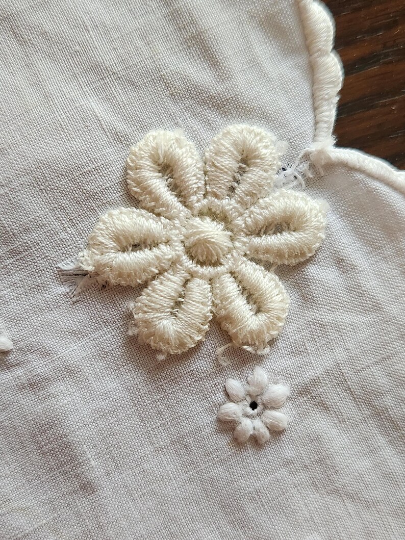 Op de afbeelding: Een wit textiel met een grote, verhoogde, cr&egrave;mekleurige geborduurde bloem met zes bloemblaadjes en een kleinere, platte, witte geborduurde bloem met een centraal oogje. De rand van de stof heeft een geschulpte, gestikte boord.