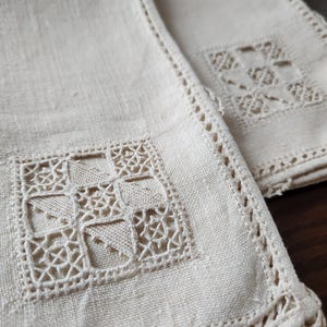 May include: Close-up of two off-white linen napkins. Each napkin features a square, openwork design in the center and a decorative, scalloped edge. The napkins are folded, showing the texture of the fabric and the intricate details of the embroidery.