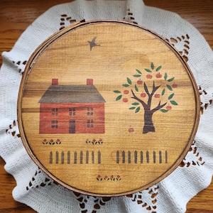 May include: Round wooden box with a painted folk art design. The design features a red house with a brown roof, a tree with red apples, a white picket fence, and a flying bird. The box is sitting on a white lace doily.