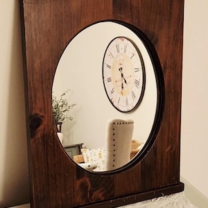 May include: A rectangular wooden framed mirror with an oval mirror. The frame is made of dark brown wood. The mirror reflects a clock and a chair.