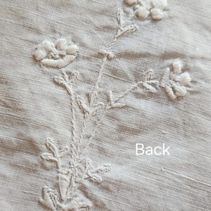 Op de afbeelding: Een lichtbeige linnen stof met witte bloemenborduursels. Het ontwerp bestaat uit een centrale stengel met bladeren en drie afzonderlijke bloemhoofdjes. Het woord Back is in witte letters gedrukt rechtsonder bij het bloemmotief.