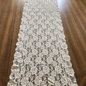 May include: A long, rectangular table runner made of off-white lace fabric. The runner features a repeating floral pattern throughout the center and a scalloped edge design along the entire perimeter, creating a delicate and intricate texture.