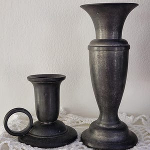 May include: Two antique pewter candlesticks. One is a short candlestick with a ring handle, and the other is a taller, vase-shaped candlestick. Both have a dark, aged finish and sit on a white lace doily.