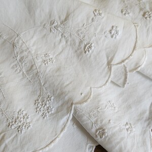 Op de afbeelding: Een wit textiel met delicate toon-op-toon bloemenborduursels met stengels, bladeren en kleine bloesems. De rand van de stof is afgewerkt met een schulprand en het borduurwerk is volledig uitgevoerd met bijpassende witte draad.