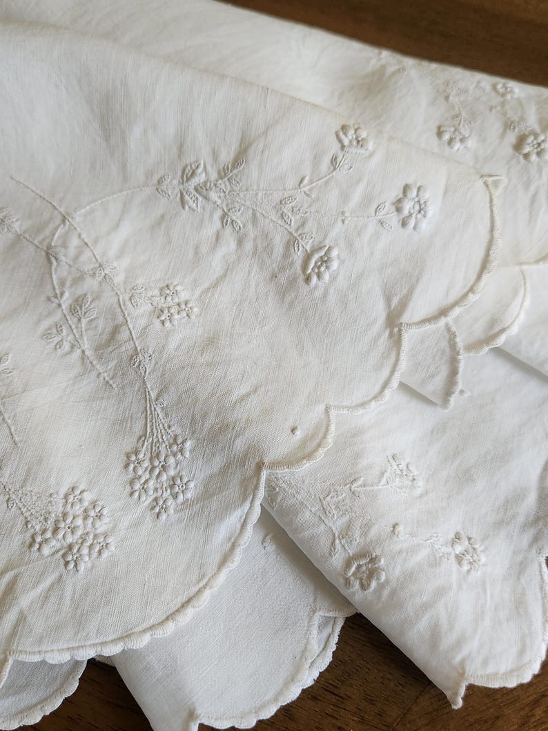 Op de afbeelding: Een wit textiel met delicate toon-op-toon bloemenborduursels met stengels, bladeren en kleine bloesems. De rand van de stof is afgewerkt met een schulprand en het borduurwerk is volledig uitgevoerd met bijpassende witte draad.