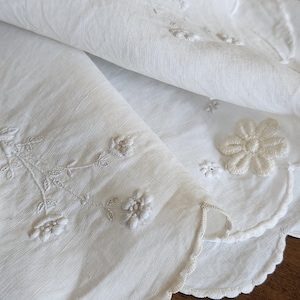 Op de afbeelding: Een wit textiel met delicate witte bloemenborduursels en geschulpte randen. Het materiaal heeft een fijne, semi-transparante textuur met gedetailleerd stikwerk van kleine bloemen en bladerige stengels langs de gebogen, afgewerkte randen.