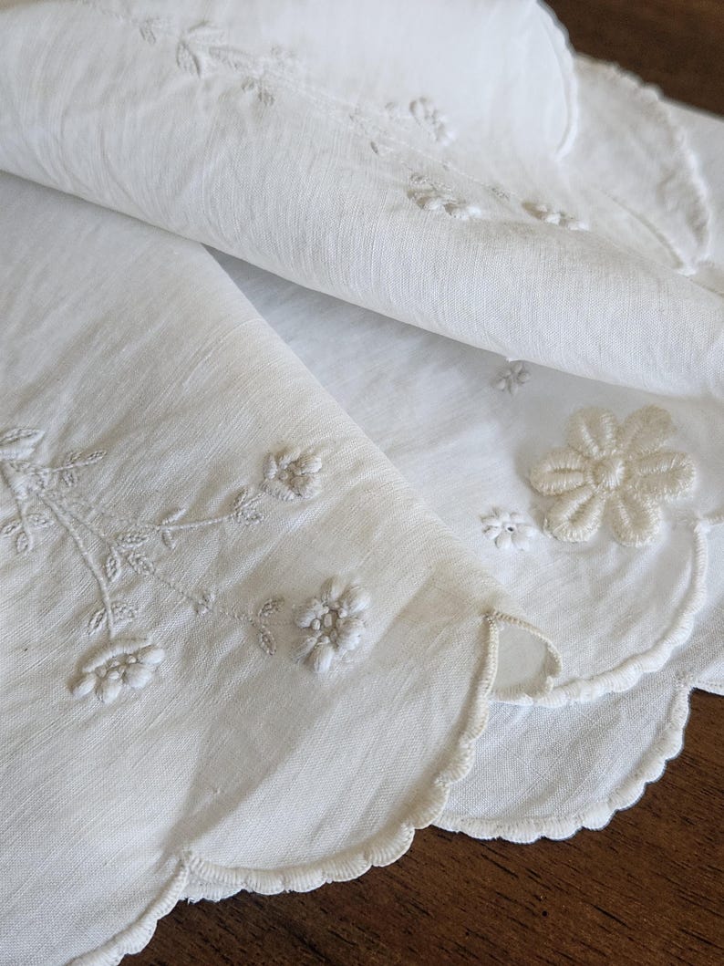 Op de afbeelding: Een wit textiel met delicate witte bloemenborduursels en geschulpte randen. Het materiaal heeft een fijne, semi-transparante textuur met gedetailleerd stikwerk van kleine bloemen en bladerige stengels langs de gebogen, afgewerkte randen.