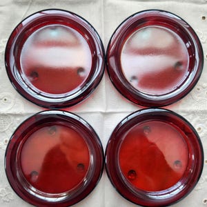 May include: Four ruby red glass plates with a circular design. Each plate has a raised rim and a slightly recessed center. The plates are arranged in a square formation, showcasing their rich color and simple elegance. The plates are likely for serving or display.