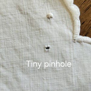Op de afbeelding: Een wit textiel met een geschulpte rand met witte borduurstiksels. De stof vertoont een klein, enkel donker gaatje nabij het midden, met de tekst Tiny pinhole in het wit gedrukt over het onderste gedeelte van de afbeelding.