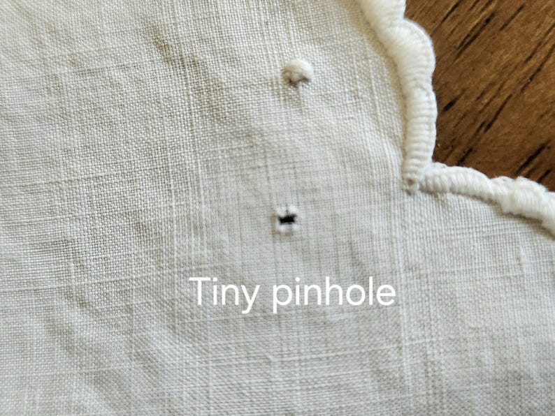 Op de afbeelding: Een wit textiel met een geschulpte rand met witte borduurstiksels. De stof vertoont een klein, enkel donker gaatje nabij het midden, met de tekst Tiny pinhole in het wit gedrukt over het onderste gedeelte van de afbeelding.