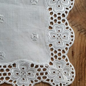 May include: Close-up of a white embroidered linen doily. The doily features a scalloped edge with floral cutouts and small embroidered flowers scattered across the surface. The background is a wooden surface.