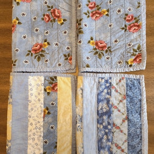 May include: Four quilted placemats with floral and patchwork designs. Two placemats feature a light blue background with red and yellow roses. The other two have vertical stripes in various floral and solid patterns, with blue and yellow hues.