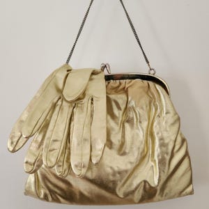 May include: A gold metallic clutch purse with a chain strap, paired with matching gloves. The purse has a kiss-lock closure and a gathered design. The gloves are draped over the purse.
