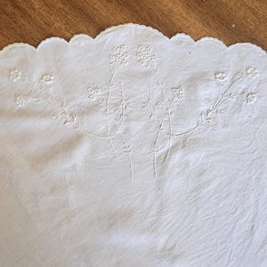 Op de afbeelding: Een wit textielstuk met verfijnd wit bloemenborduurwerk met dunne stengels en kleine bloesems. De rand van de stof is afgewerkt met een schulprand, wat een decoratieve omlijning cre&euml;ert langs de bovenste boog van het witte doek.