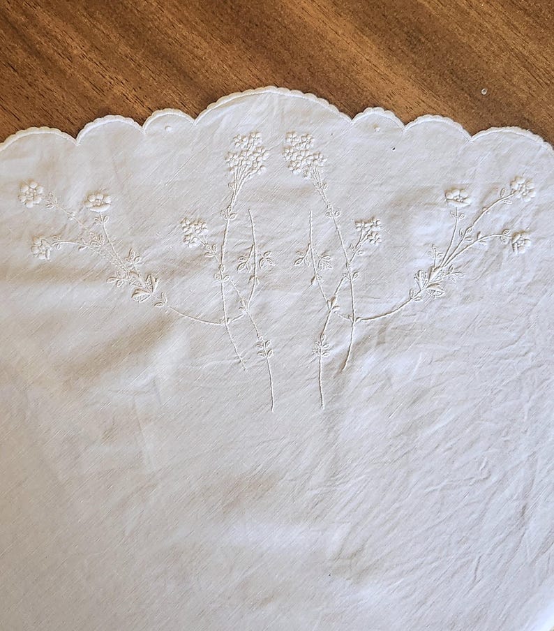 Op de afbeelding: Een wit textielstuk met verfijnd wit bloemenborduurwerk met dunne stengels en kleine bloesems. De rand van de stof is afgewerkt met een schulprand, wat een decoratieve omlijning cre&euml;ert langs de bovenste boog van het witte doek.
