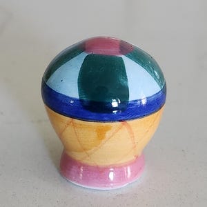 May include: A small, rounded ceramic knob featuring a hand-painted design. The top section displays alternating segments of dark green, light blue, and pink. A horizontal blue band separates the top from the yellow base, which has a cross-hatched pattern.