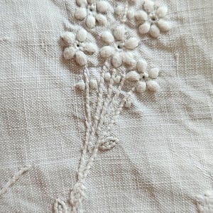 Op de afbeelding: Een lichtbeige stof met witte bloemenborduursels. Het ontwerp bestaat uit een trosje kleine, verhoogde bloemen met vijf bloemblaadjes, inclusief gestikte stengels en blaadjes. Het borduurwerk maakt gebruik van de satijnsteektechniek, wat zorgt voor een textuurrijk en ruimtelijk uiterlijk op het geweven textieloppervlak.