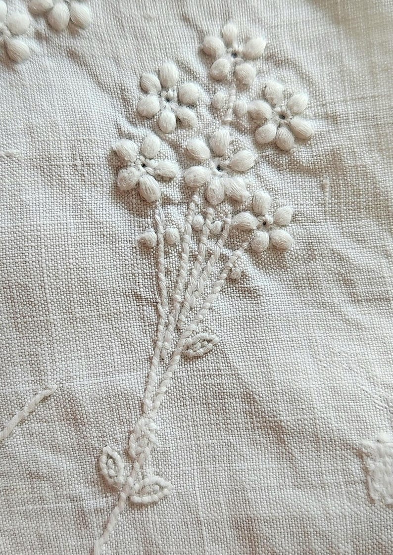 Op de afbeelding: Een lichtbeige stof met witte bloemenborduursels. Het ontwerp bestaat uit een trosje kleine, verhoogde bloemen met vijf bloemblaadjes, inclusief gestikte stengels en blaadjes. Het borduurwerk maakt gebruik van de satijnsteektechniek, wat zorgt voor een textuurrijk en ruimtelijk uiterlijk op het geweven textieloppervlak.