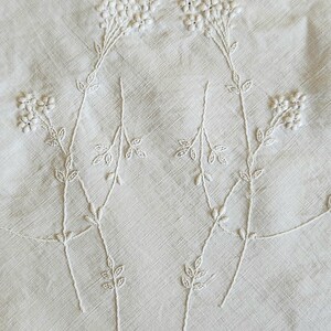Op de afbeelding: Een wit linnen textiel met delicate witte bloemenborduursels. Het ontwerp toont twee hoofdstelen met trossen kleine bloemen en bladeren, uitgevoerd in ton-sur-ton draadwerk. De rand van de stof is afgewerkt met een geschulpte boord.