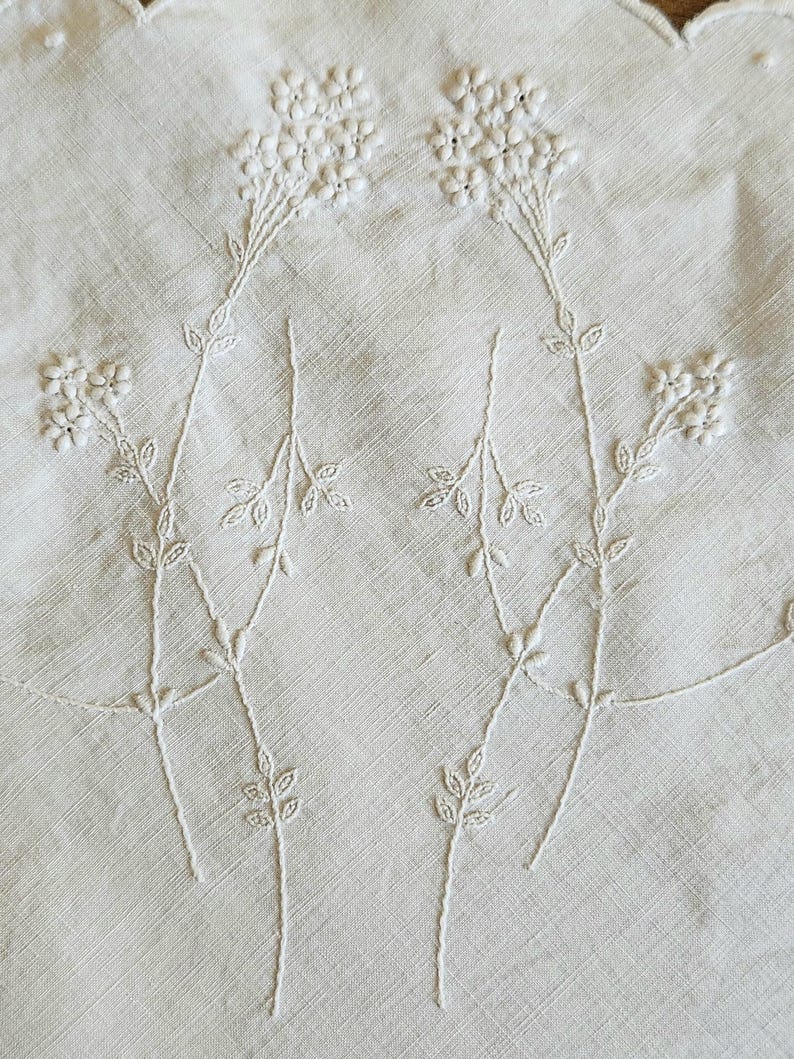 Op de afbeelding: Een wit linnen textiel met delicate witte bloemenborduursels. Het ontwerp toont twee hoofdstelen met trossen kleine bloemen en bladeren, uitgevoerd in ton-sur-ton draadwerk. De rand van de stof is afgewerkt met een geschulpte boord.