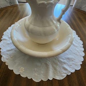 Op de afbeelding: Een waskom en kan set van wit keramiek met een geschulpte rand en een gebogen handvat. De kan staat in de brede, ondiepe kom, beide rustend op een wit rond stoffen kleedje met geborduurde bloemendetails.