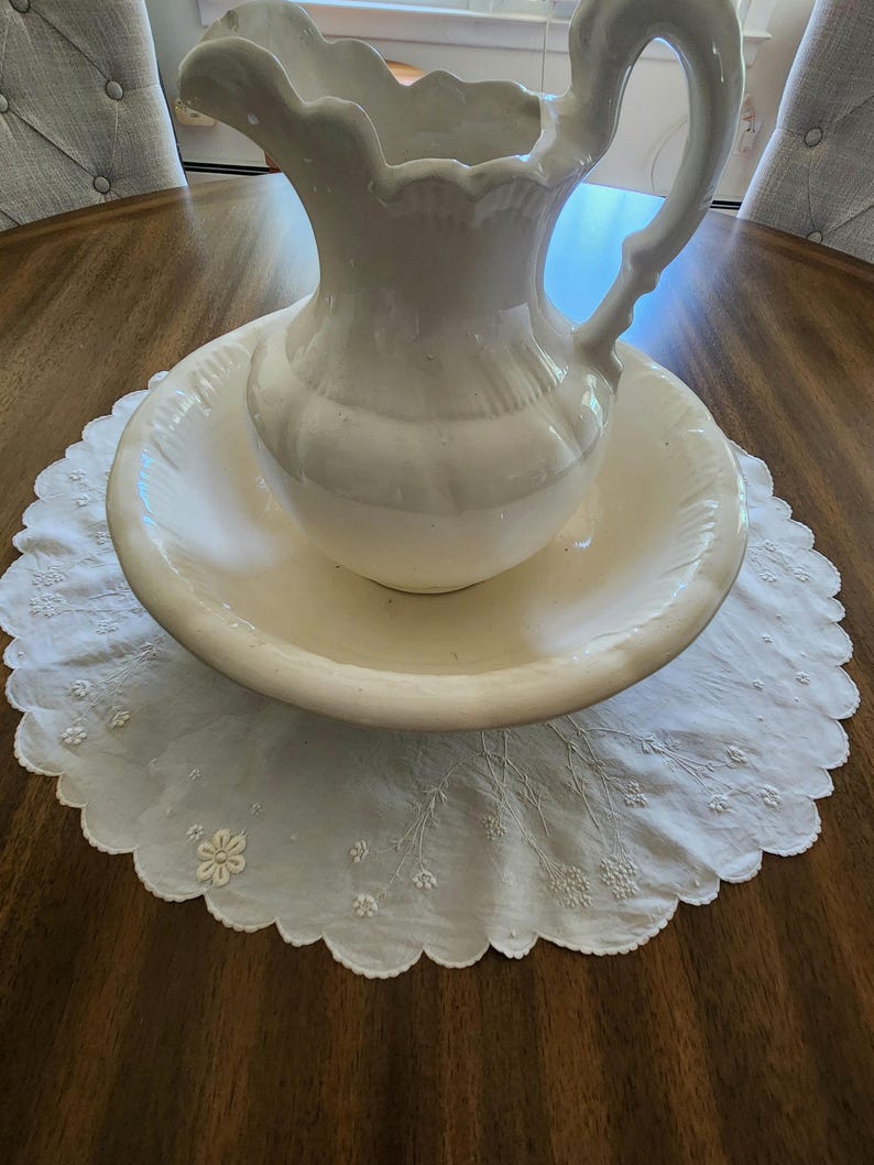 Op de afbeelding: Een waskom en kan set van wit keramiek met een geschulpte rand en een gebogen handvat. De kan staat in de brede, ondiepe kom, beide rustend op een wit rond stoffen kleedje met geborduurde bloemendetails.
