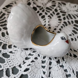 May include: A white ceramic dove-shaped planter with a gold rim around the opening. The dove has a red beak and black eyes. The planter is sitting on a white lace doily.