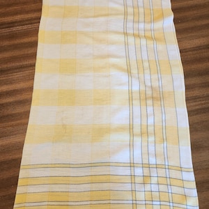 May include: A rectangular kitchen towel featuring a yellow and white checkered pattern. The fabric displays a grid of soft yellow squares interspersed with white sections, accented by thin, dark grey vertical and horizontal pinstripes.